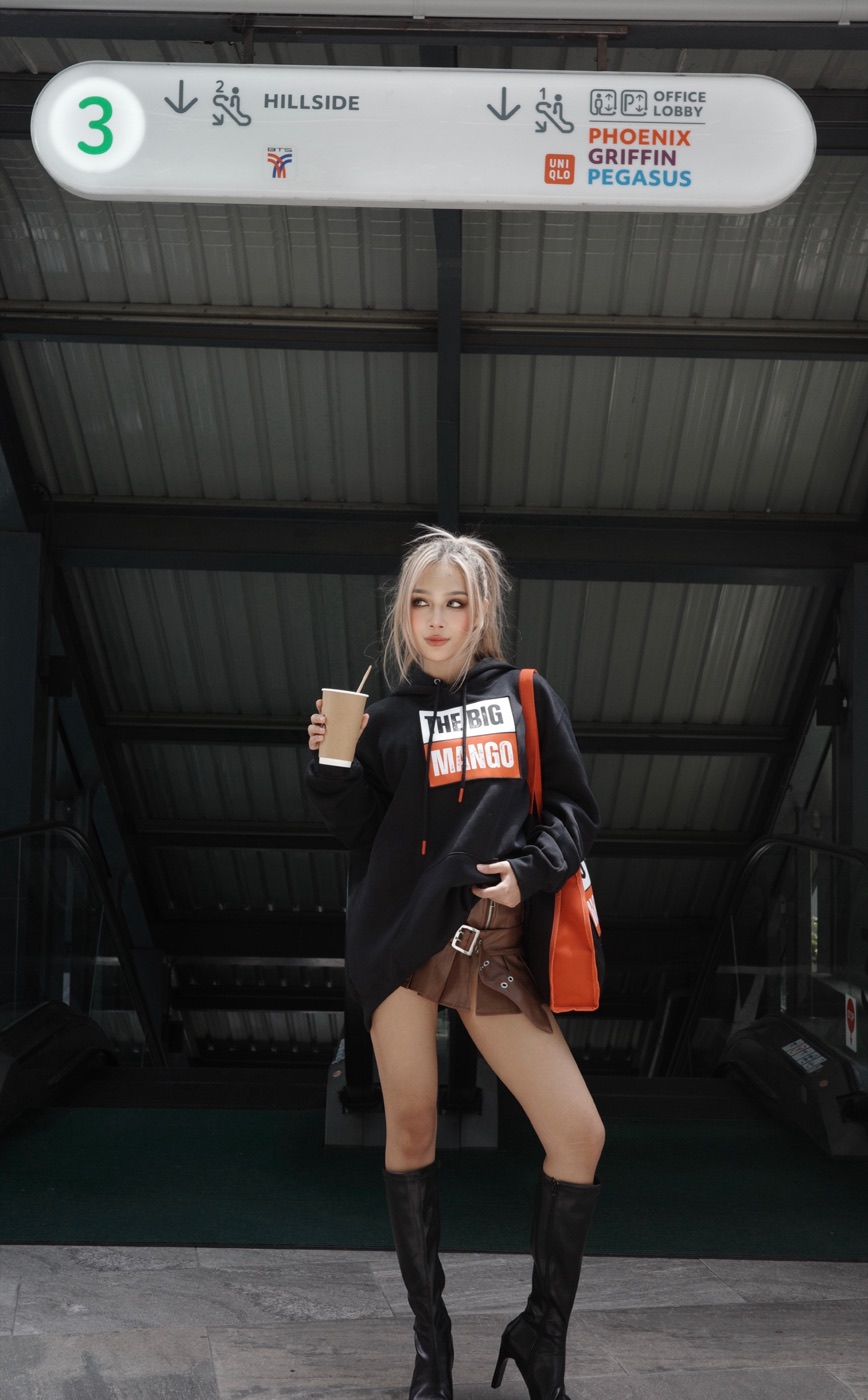 Model in the The Big Mango hoodie holding an iced coffee at a Bangkok BTS Skytrain station