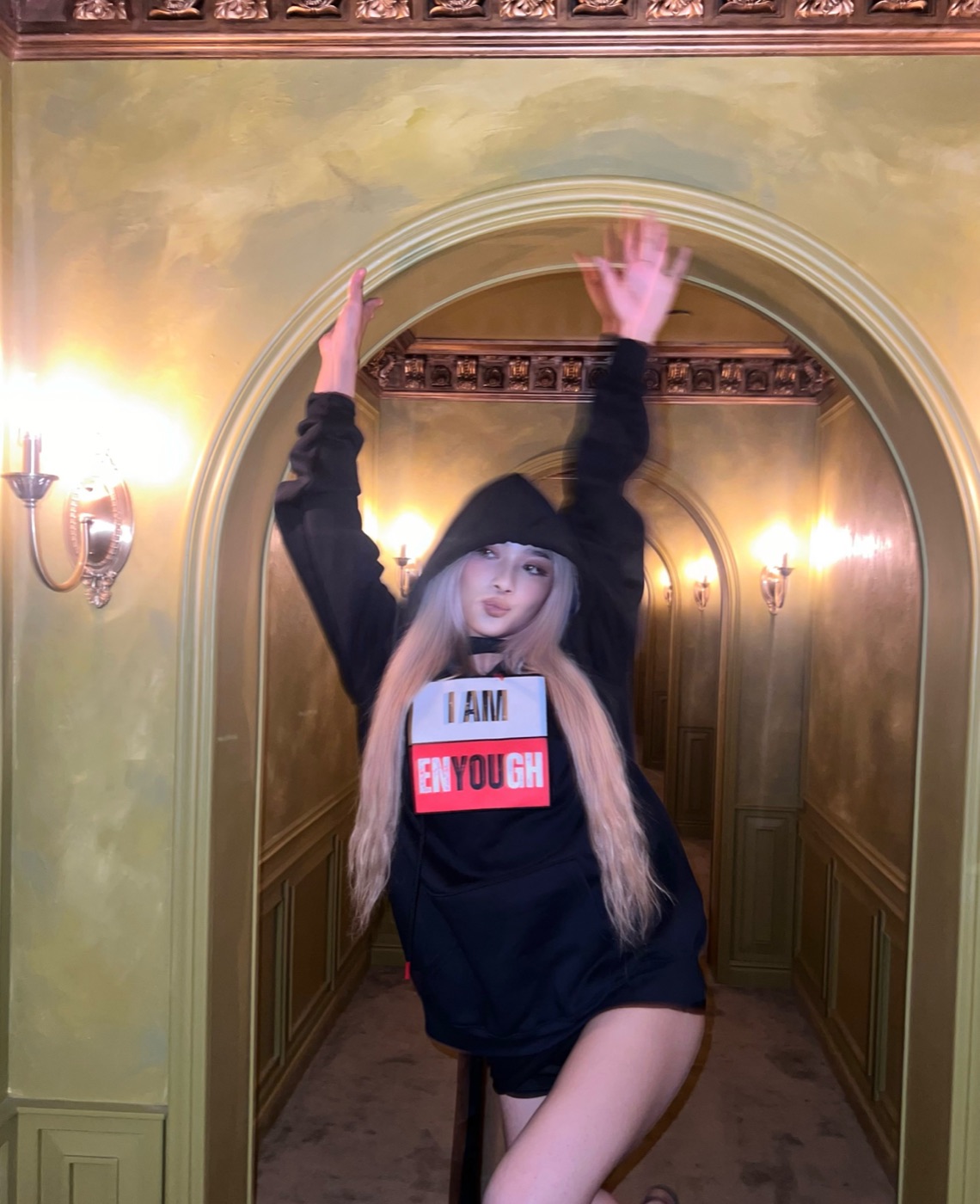 Model in the I Am Enyough hoodie, mid-motion with arms raised in an ornate gold hallway