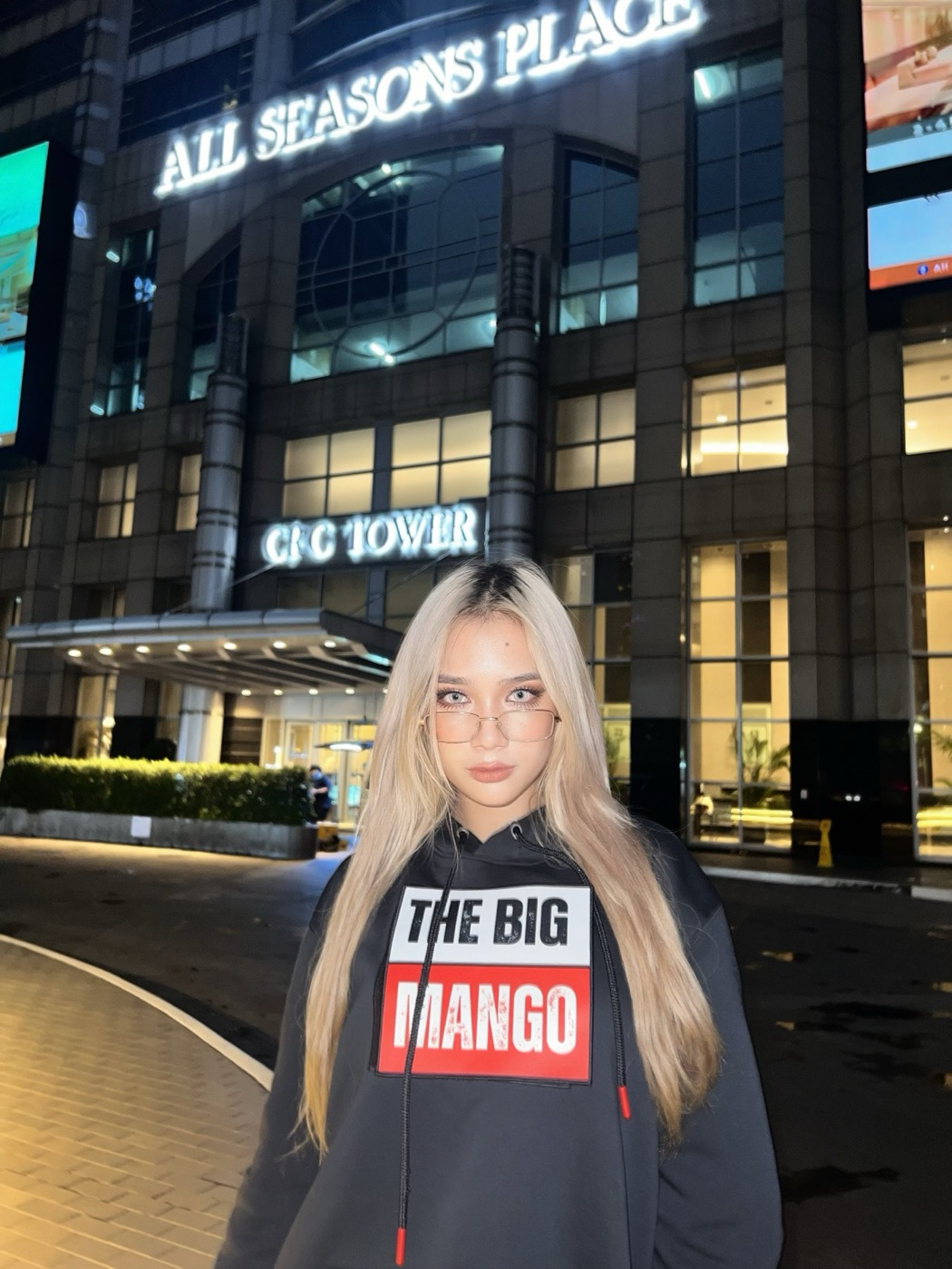 Model in the The Big Mango hoodie on a Bangkok street at night, illuminated storefront signs behind her