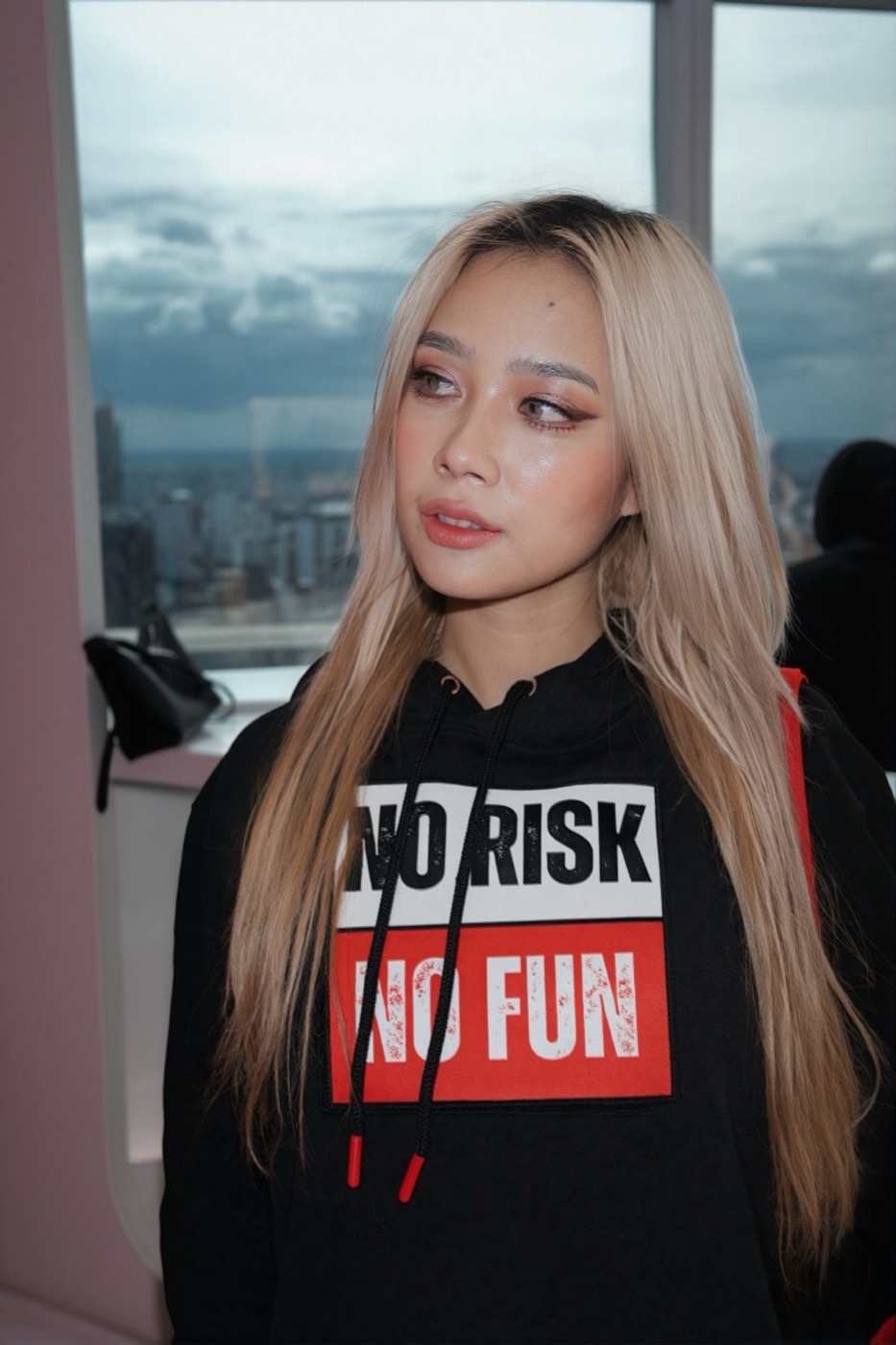 Candid portrait of the model in the No Risk No Fun hoodie, Bangkok skyline through the window behind her