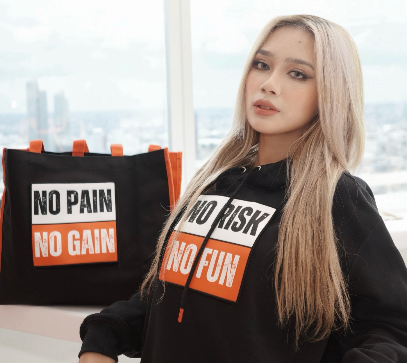 Close-up of the model in the No Risk No Fun hoodie beside the No Pain No Gain Signal Tote, rooftop