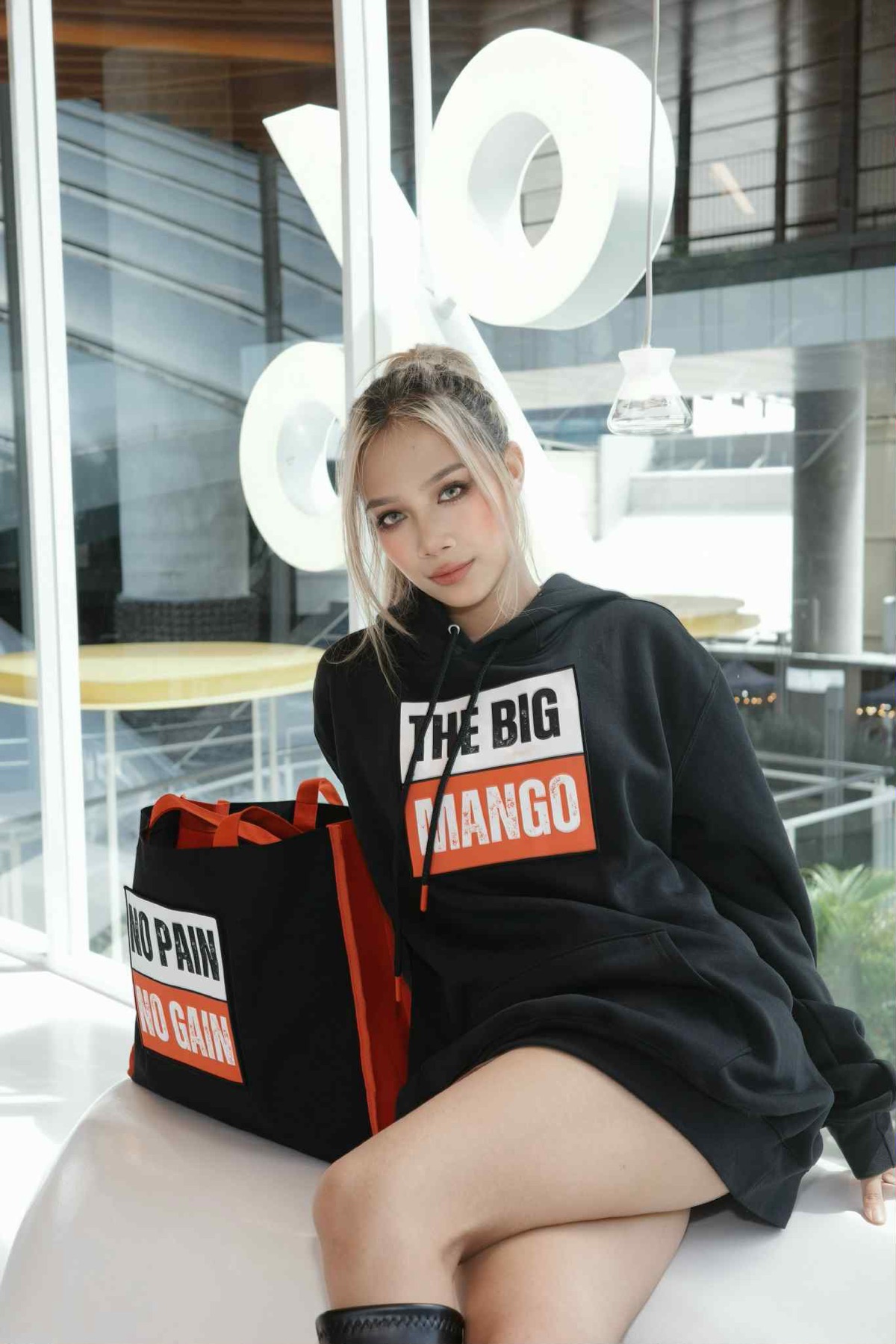 Model wearing The Big Mango hoodie, seated in front of a glass building
