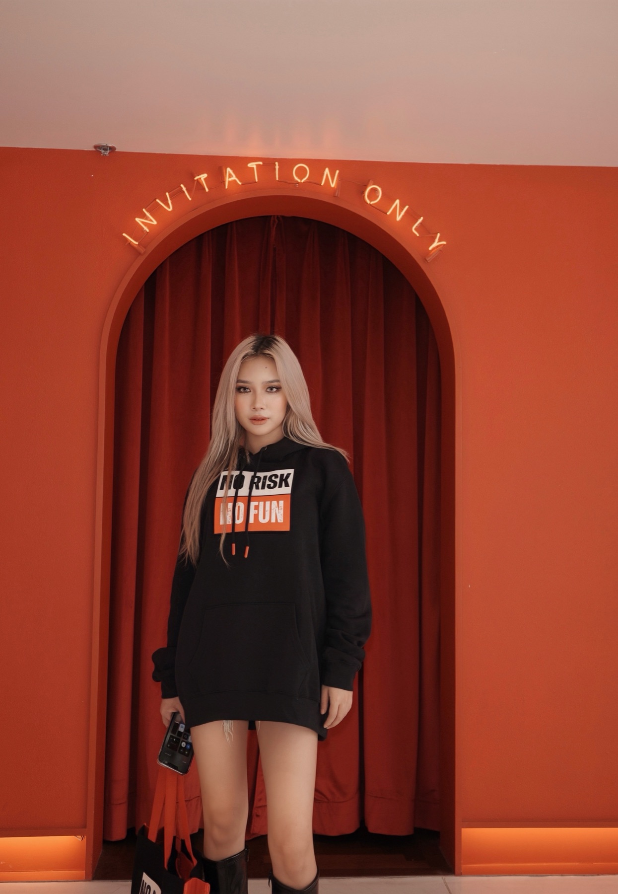 ENYOUGH model in the No Risk No Fun hoodie under a neon 'Invitation Only' sign at a Bangkok lounge